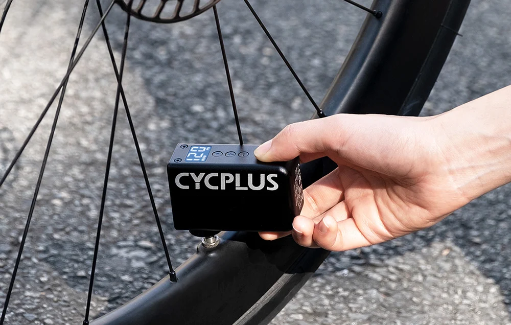 Dłoń rowerzysty trzymająca precyzyjną pompkę elektryczną Cycplus AS2 PRO podłączoną do wentyla opony szosowej.